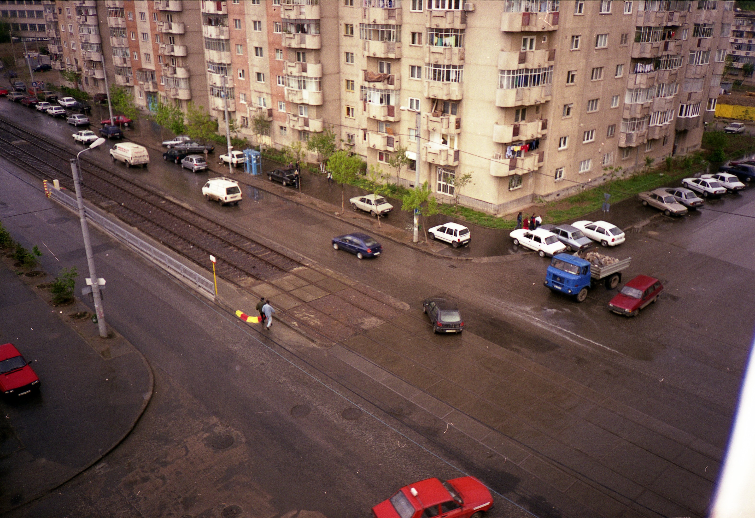 Luftaufnahme einer Stadtstraße mit parkenden Autos, Gebäuden, Bäumen, Laternen, Bahnschienen und Fußgängern, mit Text oben und unten.
