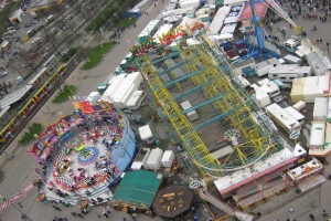 Luftaufnahme eines Vergnügungsparks mit einem zentralen Riesenrad, umgeben von Fahrgeschäften, Hütten, Fahrzeugen, Menschen, Straßen, Pfählen, Bäumen und Gras.