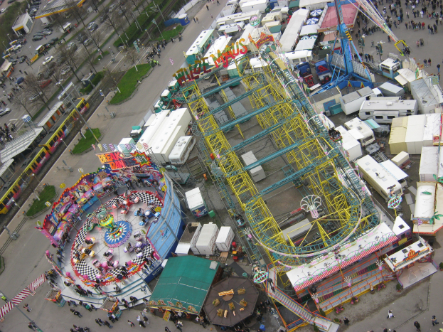 Luftaufnahme eines Vergnügungsparks mit einem zentralen Riesenrad, umgeben von Fahrgeschäften, Hütten, Fahrzeugen, Menschen, Straßen, Pfählen, Bäumen und Gras.