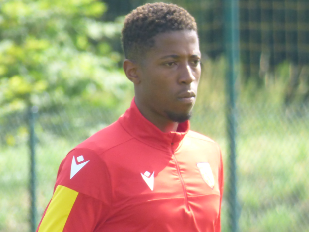 Ein junger Mann in einem roten und gelben T-Shirt steht auf einem Fußballfeld umgeben von einem Zaun und Bäumen und schaut entschlossen während eines Trainings.