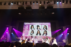 Eine Gruppe von Menschen auf einer hell erleuchteten Bühne mit Trägern, wobei einer ein Mikrofon hält und ein anderer ein Instrument spielt, während eine Menge mit Kameras unten zusieht und ein großes Display Bilder der K-Pop-Gruppe BK48 zeigt.