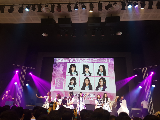 Eine Gruppe von Menschen auf einer hell erleuchteten Bühne mit Trägern, wobei einer ein Mikrofon hält und ein anderer ein Instrument spielt, während eine Menge mit Kameras unten zusieht und ein großes Display Bilder der K-Pop-Gruppe BK48 zeigt.