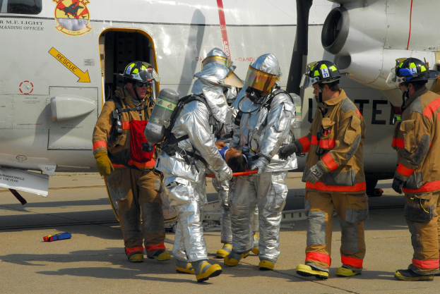 Eine Gruppe von Feuerwehrleuten in Helmen und Schutzausrüstung steht auf einer Flugzeuglandebahn, einer hält ein Objekt, mit verschiedenen Gegenständen drumherum verstreut.