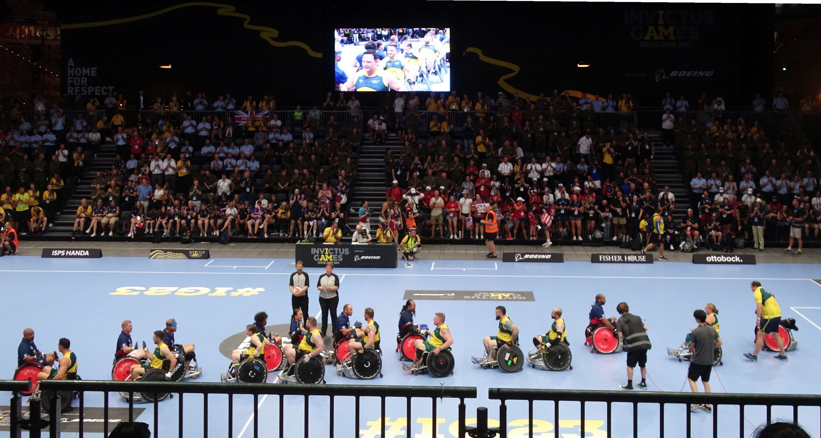 Eine Gruppe von Rollstuhlfahrern bei den Invictus Games Düsseldorf 2020 auf einem von einem Geländer umgebenen Platz, mit einer großen Menge im Hintergrund und einer Treppe, die zu einem Bildschirm führt.