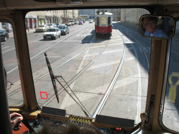 Ein Mann fährt eine Tram auf einer Stadtstraße mit anderen Fahrzeugen, Gebäuden und einem klaren blauen Himmel im Hintergrund.