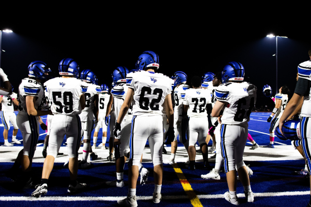 Eine Gruppe von Fußballspielern in Helmen und Fußballschuhen steht auf einem Feld, mit beleuchteten Pfosten im Hintergrund.