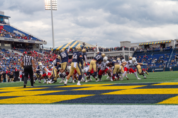 Ein Football-Spiel im Gange mit Spielern in Sportbekleidung und Helmen im Vordergrund, Zuschauern, Geländern, Tafeln mit Text, Laternen und einem bewölkten Himmel im Hintergrund und Text am unteren Bildrand.