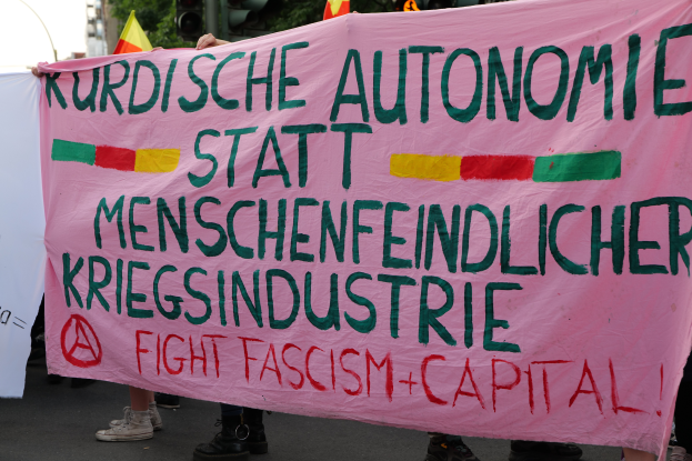 Eine Gruppe von Menschen steht auf einer Straße und hält ein pinkes Banner mit der Aufschrift "Kurdische Autonomie Statt Menschenfeindlicher Kriegsindustrie Fight Fascism Capital". Im Hintergrund sind Verkehrsschilder, Flaggen, Bäume, Gebäude und ein klarer blauer Himmel zu sehen.
