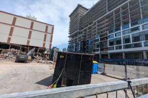Großes Gebäude mit Schutt davor, umgeben von einem Metallzaun, einem Container, Pflanzen, Bäumen, Pfählen, einem Schild, Fahrzeugen, Steinen und einem bewölkten Himmel an einer Baustelle für einen neuen Wohnkomplex.