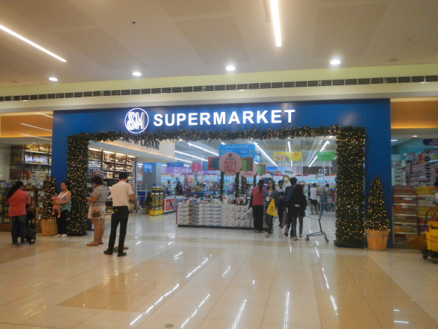 Gruppe von Menschen vor einem Supermarkt mit Einkaufstägern, Taschen und einem Blumentopf, Regalen, Schildern und Deckenbeleuchtung.