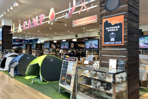 Ein Geschäft voller Campingausrüstung, darunter Zelte, Schilder, eine Glasvitrine, Fernseher, Ständer mit Gegenständen, Deckenleuchten und andere Objekte.