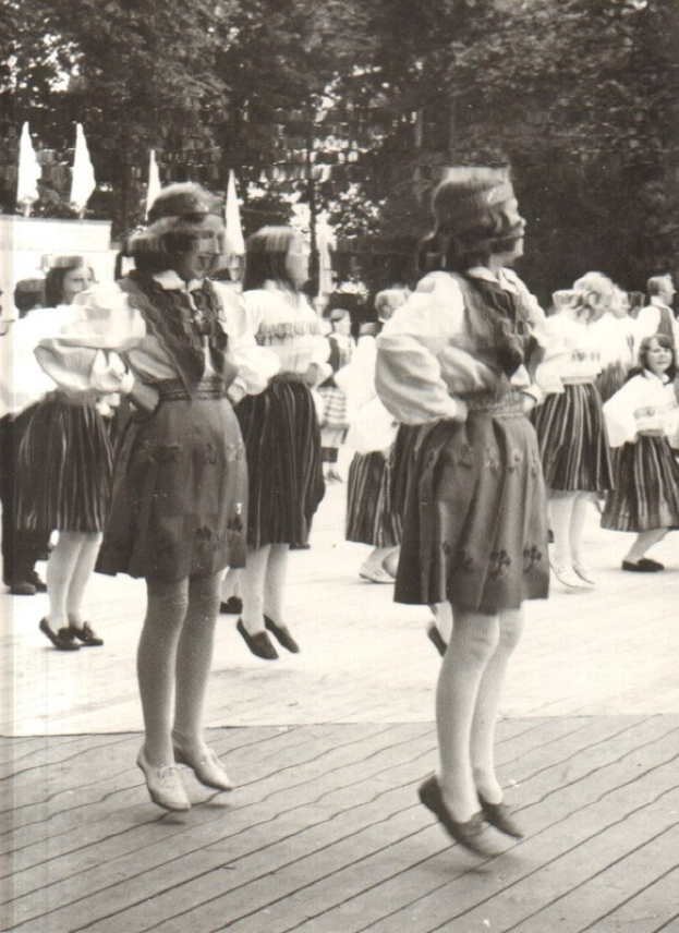 Schwarzes und weißes Foto von Frauen in traditioneller bayrischer Tracht, die auf einem hölzernen Boden umgeben von Bäumen und Fahnen tanzen.