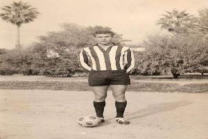 Ein Schwarz-Weiß-Foto eines älteren Mannes, der einen Fußball hält und vor Bäumen und Himmel steht.