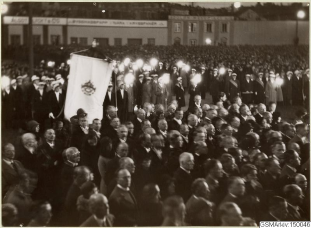 Schwarzes und weißes Foto einer großen Menschenmenge vor einem Gebäude, mit einer Person, die eine Fahne hält, in der Mitte, Pfosten und Lichter im Hintergrund und Text unten, der lautet: "Gustav Klimts Rede an der Berliner Mauer."