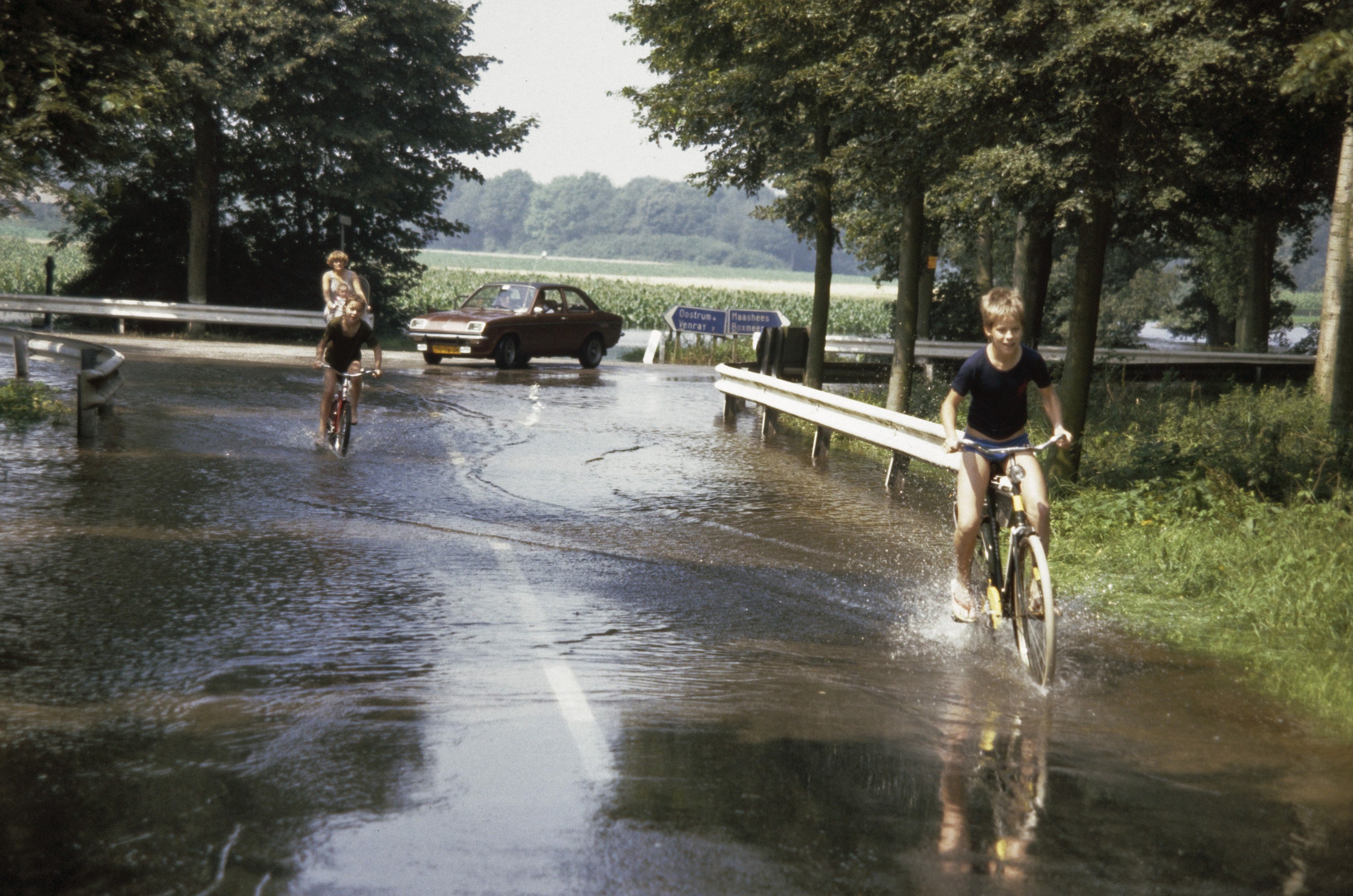 Eine Gruppe von Menschen, die auf Fahrrädern eine überflutete Straße entlangfahren, mit einem danebenfahrenden Auto, gesäumt von Geländern, Bäumen und Pflanzen unter einem klaren blauen Himmel.