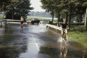 Eine Gruppe von Menschen, die auf Fahrrädern eine überflutete Straße entlangfahren, mit einem danebenfahrenden Auto, gesäumt von Geländern, Bäumen und Pflanzen unter einem klaren blauen Himmel.
