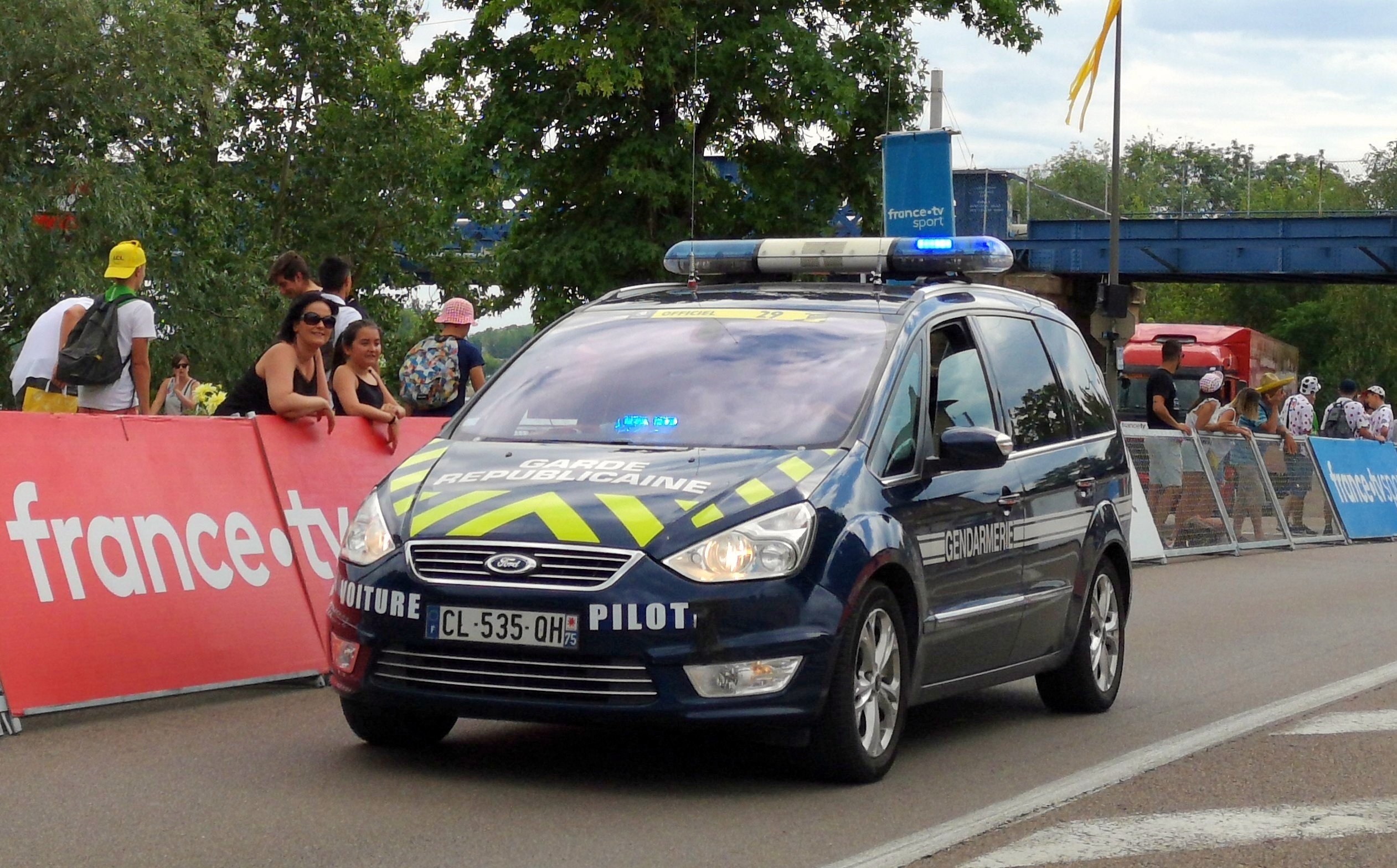 Polizeiauto fährt an einer Menge mit Schildern, Geländern, Bäumen, einer Brücke, einer Flagge und einem bewölkten Himmel vorbei.
