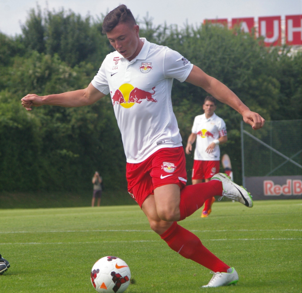 Ein professioneller Fußballspieler schießt einen Fußball auf einem grünen Feld mit Bäumen und einem klaren blauen Himmel im Hintergrund, wobei "RB Leipzig" auf einer Tafel zu sehen ist.