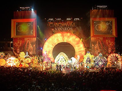Große Menschenmenge sammelt sich nachts vor einer hell erleuchteten Bühne mit der Aufschrift "Carnaval de Vinaros", was auf ein Karnevals-Event hindeutet.