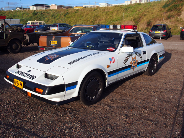 Ein Polizeiwagen auf einem Parkplatz neben anderen Autos, Gras, Pfosten, Drähte, Gebäude und einen klaren blauen Himmel.