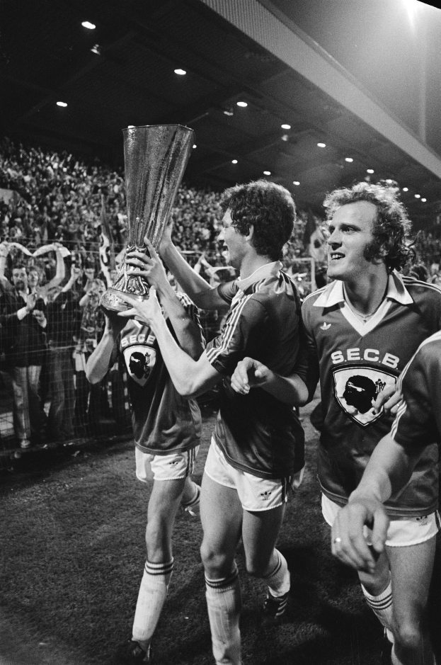 Schwarzes und weißes Bild von Männern, die einen Fußballsieg feiern, indem sie einen Pokal in die Höhe halten, auf einem Feld, mit einer Menge hinter einem Zaun und überdachter Stadionbeleuchtung.