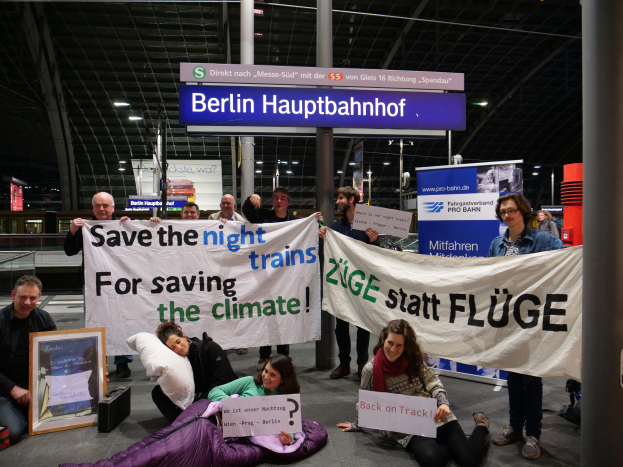 Eine Gruppe von Menschen sitzt auf dem Boden des Berliner Hauptbahnhofs und hält ein Banner mit der Aufschrift "Retten Sie die Nachtzüge, um das Klima zu retten". Sie sind von Kissen, einem Foto Rahmen und anderen Gegenständen umgeben. Im Hintergrund sind Bretter, Säulen, Lampen und Geländer zu sehen.