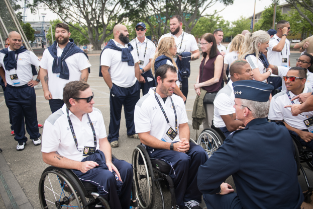Eine Gruppe von Menschen im Rollstuhl, die vor einer Kulisse aus Bäumen, Pfählen, Gebäuden und einem klaren blauen Himmel sprechen, wobei einige Mützen und Brillen tragen, was darauf hindeutet, dass sie bei den Paralympischen Spielen sind.