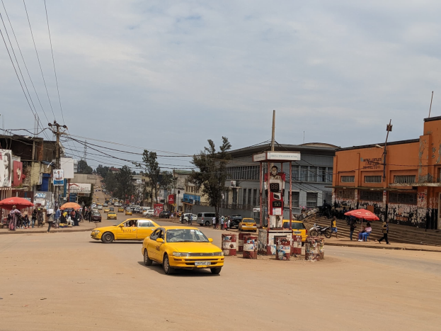 Ein gelbes Taxi fährt eine belebte Stadtstraße entlang, die von Gebäuden gesäumt ist, während Fußgänger mit Regenschirmen auf dem Gehweg darunter gehen, unter bewölktem Himmel.