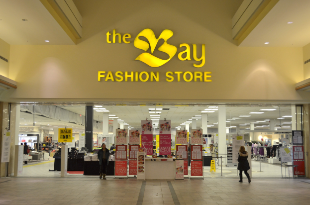 Außenansicht eines Bay Fashion Stores in einem Einkaufszentrum mit Menschen, Texttafeln an den Wänden, Säulen, Deckenleuchten und Hintergrundanzeigen.