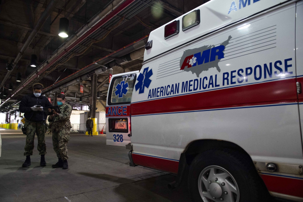 Notarztwagen der American Medical Response drinnen geparkt mit zwei maskierten Personen davor, ein Fahrzeug links und zusätzliche Personen und Gegenstände im Hintergrund.