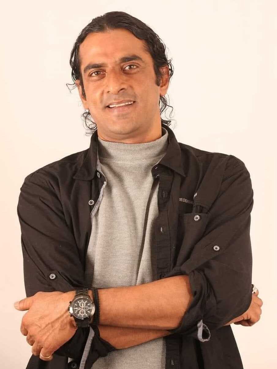 Ein Mann in einem schwarzen und aschfarbenen Kleid mit überschlagenen Armen, der eine Uhr trägt und gegen einen weißen Hintergrund steht, der tief nachdenklich erscheint.