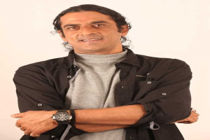 Ein Mann in einem schwarzen und aschfarbenen Kleid mit überschlagenen Armen, der eine Uhr trägt und gegen einen weißen Hintergrund steht, der tief nachdenklich erscheint.