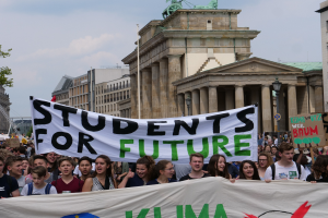 Gruppe von Schülern marschiert in Berlin mit einer bunt bemalten "Students for Future"-Schultertasche vor einem Hintergrund aus Gebäuden, Bäumen und Himmel.