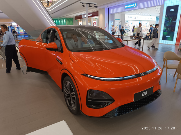 Ein oranges Elektroauto in einer Ausstellungsreihe umgeben von Menschen, mit Stühlen und einem Wagen rechts und Geschäften im Hintergrund.