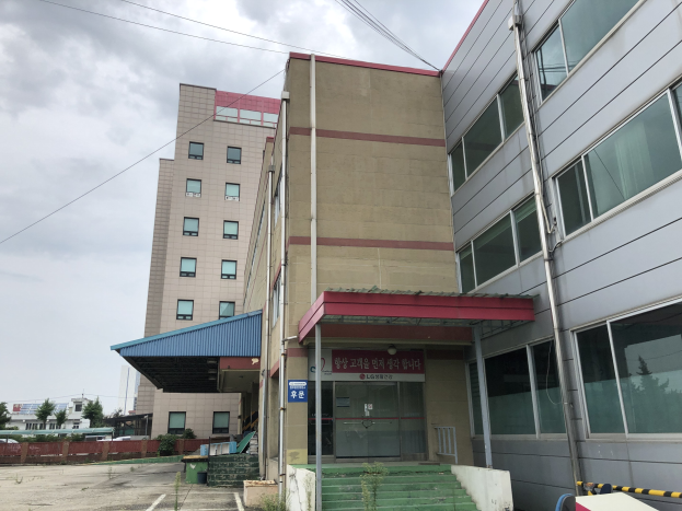 Großes Gebäude mit vielen Fenstern, als Hauptquartier der Koreanischen Nationalen Universität für Wissenschaft und Technologie bezeichnet, umgeben von Grünflächen, einer Treppe, einem Schild, Pfählen, einem Zaun, Bäumen, Fahrzeugen, Drähten und einem bewölkten Himmel.