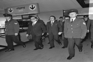 Schwarzes und weißes Foto von uniformierten Männern, die durch einen Flughafen gehen, mit einer Tafel an der Wand im Hintergrund.