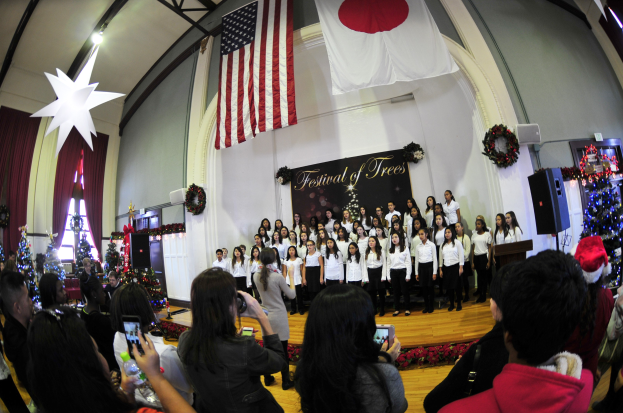 Eine Gruppe von Menschen auf einer Bühne vor einem Publikum mit Weihnachtsbäumen, einem Rednerpult, Lautsprechern, einer Texttafel, Vorhängen, Flaggen und Deckenleuchten.