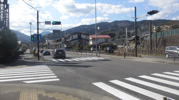Eine Stadtstraße mit Autos, einer Fußgängerfurt, Strommasten, Ampeln, Gebäuden, Bäumen, Hügeln und einem bewölkten Himmel.