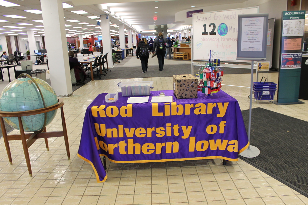 Eine Expo-Szene mit einem Tisch, auf dem verschiedene Landesflaggen, eine Box, Papiere und ein Banner sowie links daneben eine Weltkugel und rechts daneben eine Anschlagtafel mit einigen Menschen und im Hintergrund viele Tische und Stühle zu sehen sind.