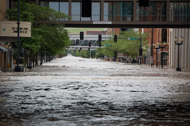 Überflutete Stadtstraße mit Wasser, das die Straße, Pfosten, Laternen, Schilder, Verkehrszeichen, Bäume, Gebäude und eine Brücke im Hintergrund bedeckt.