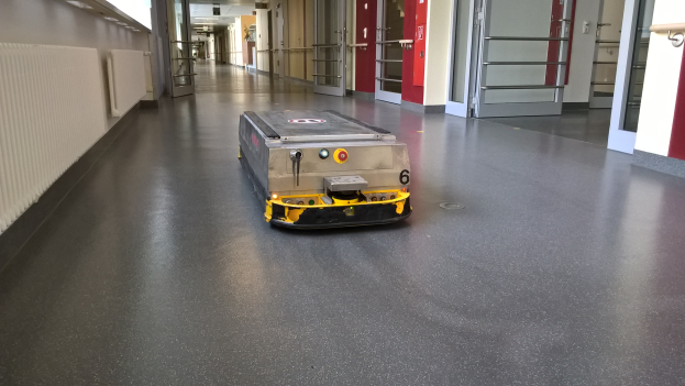 Ein Roboter, wahrscheinlich ein Reinigungsgerät, steht auf dem Boden eines Gebäudes, umgeben von Wänden, Türen, Lampen und anderen Gegenständen.