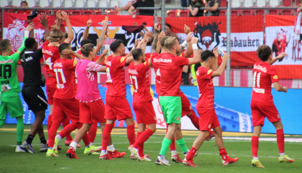 Gruppe von Fußballspielern, die ein Tor feiern, auf einem Feld, einige halten Flaschen oder klatschen, mit Bannern, einem Zaun, Metallstangen, Stühlen und einem Dach mit Lampen im Hintergrund.