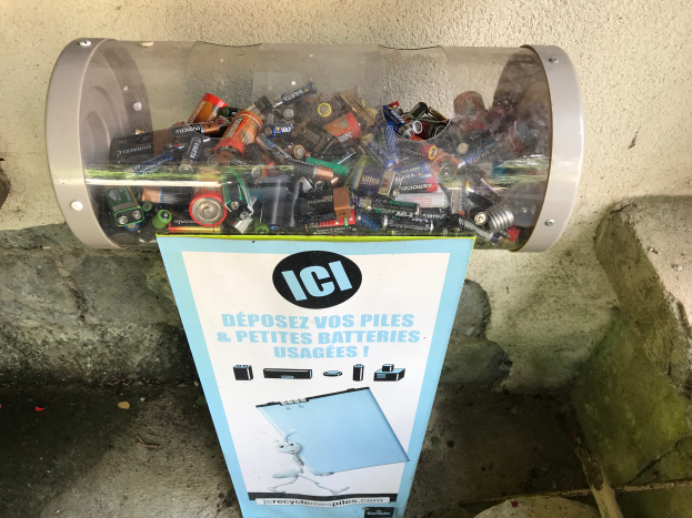 Ein Mülltonnen voller Batterien auf einem Bürgersteig, mit einem Plakat mit Text an der Wand dahinter und Treppen auf der rechten Seite.