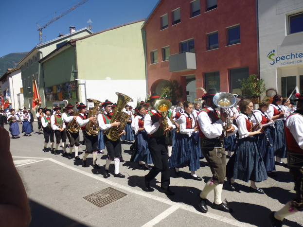 Eine Gruppe von Menschen in traditioneller bayrischer Tracht marschiert auf einer Straße, spielt Instrumente und trägt Fahnen, mit Gebäuden, Grünfläche und einem Hügel im Hintergrund unter einem klaren Himmel.