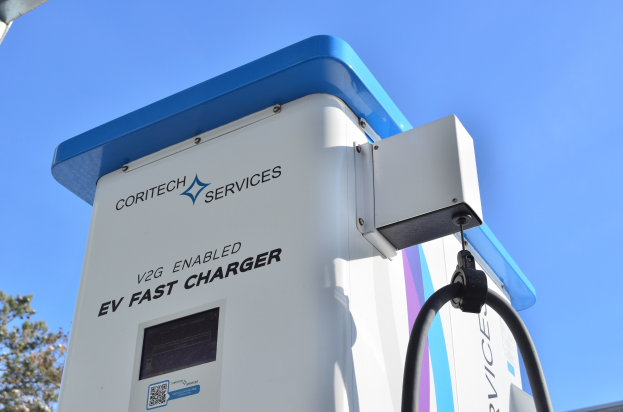 Elektrisches Fahrzeug-Ladestation mit einem "V2G Fast Charger"-Schild, umgeben von Bäumen und einem Gebäude unter einem klaren blauen Himmel.